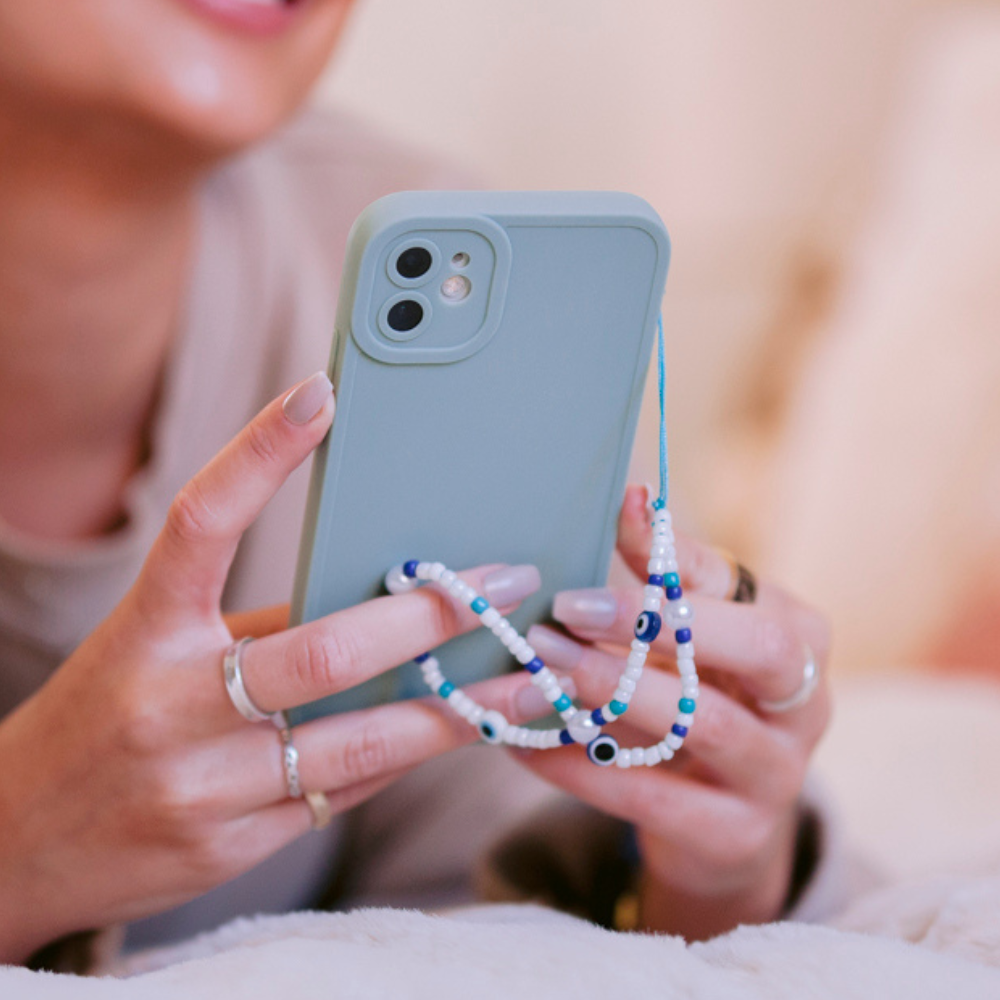 A person is holding a light blue smartphone adorned with a beaded phone charm and encased in the NAKD Phone Case - Baby Blue by Coconut Lane, which provides full camera protection. Their manicured nails and multiple rings add to the stylish look. The focus remains on the phone, while the person's face is blurred in the background.