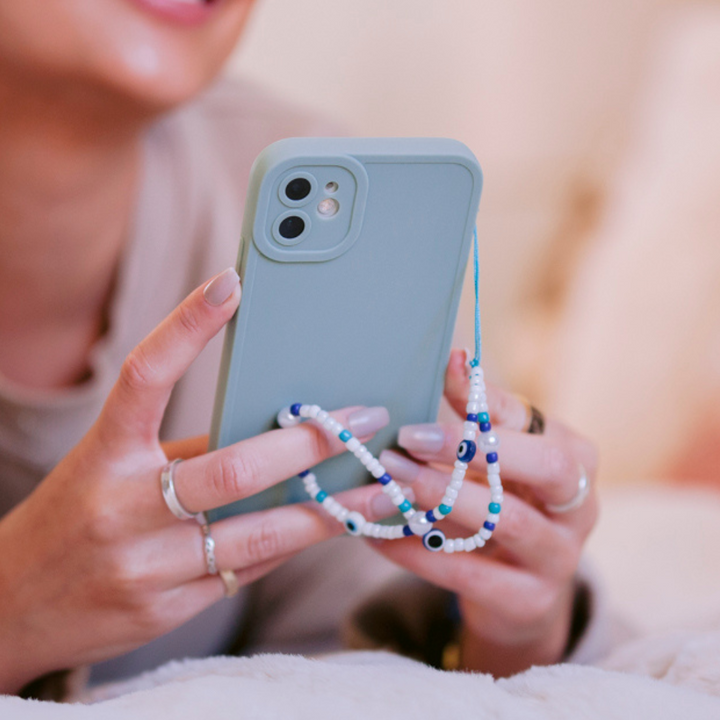 A person is holding a light blue smartphone adorned with a beaded phone charm and encased in the NAKD Phone Case - Baby Blue by Coconut Lane, which provides full camera protection. Their manicured nails and multiple rings add to the stylish look. The focus remains on the phone, while the person's face is blurred in the background.