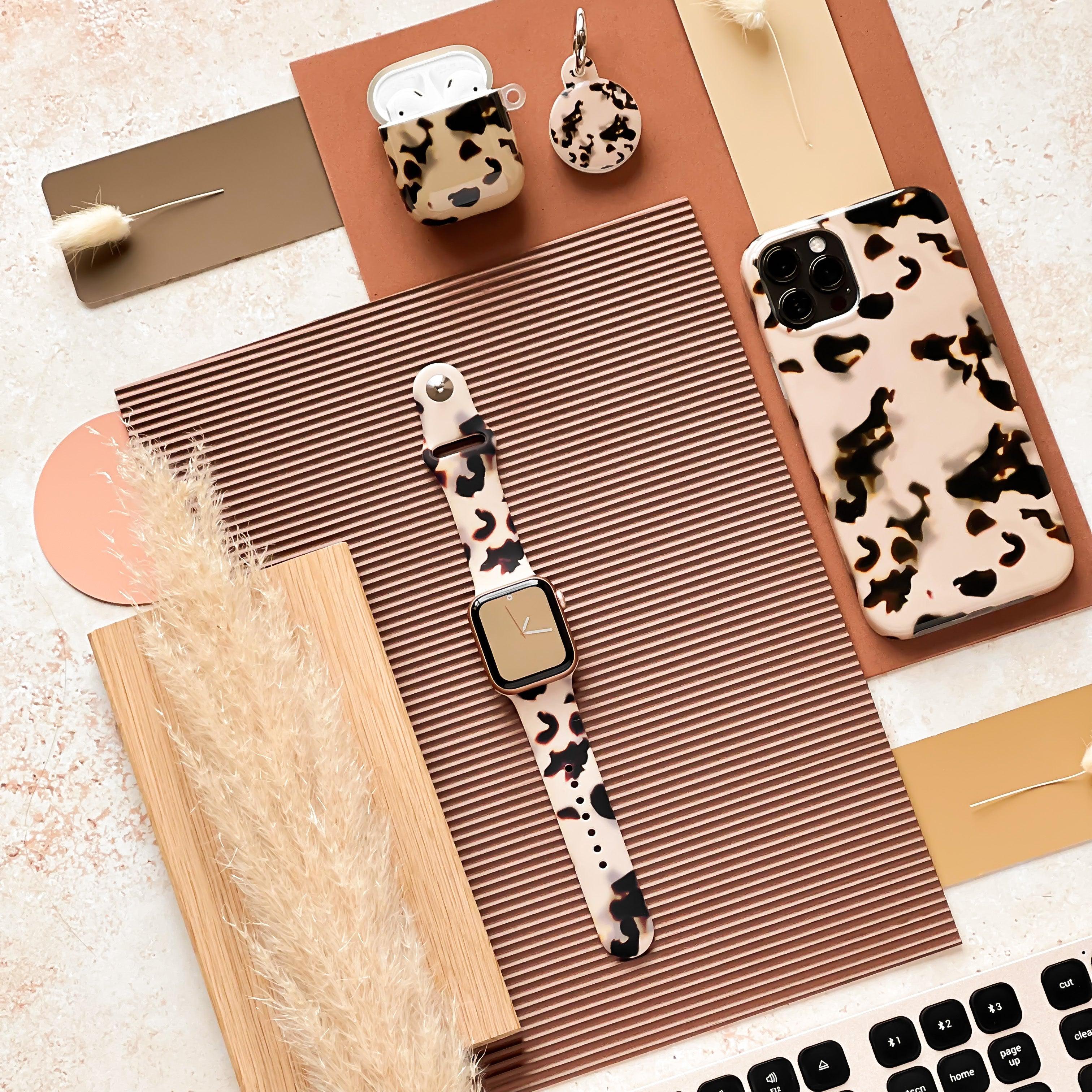 A flat lay photo showcases a stylish collection of tech accessories, including a leopard print smartwatch with a Milky Ivory Tort Apple Watch Strap from Coconut Lane, a matching phone case, an AirPods case, and a keychain. The items are arranged on textured surfaces with decorative dried plants, creating an aesthetically pleasing scene.
