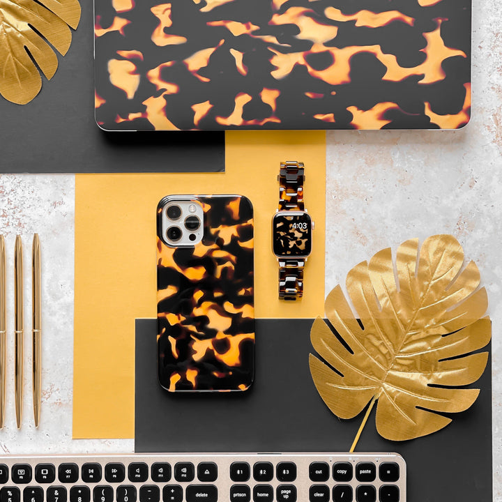A flat lay features a smartphone, a smartwatch adorned with the Luxe Tortoiseshell Apple Watch Strap by Coconut Lane, and a laptop showcasing matching orange and black swirled patterns. Arranged on a yellow and grey surface, the setup includes gold pens, a black keyboard, and gold-painted tropical leaves for an added aesthetic touch.