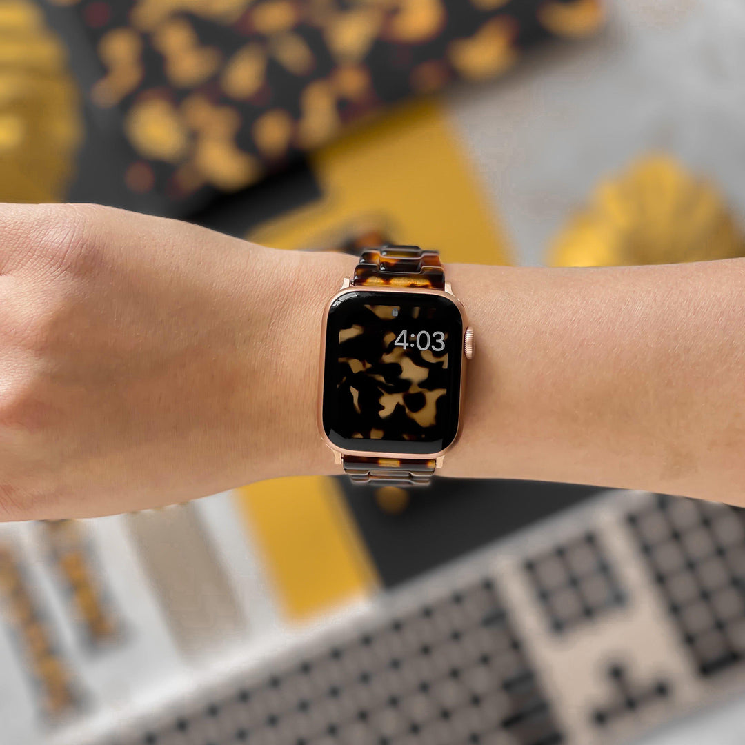 A close-up photo shows a person wearing a gold-colored Apple Watch paired with Coconut Lane's Luxe Tortoiseshell Apple Watch Strap, featuring adjustable shades of brown and yellow. The watch face displays the time "4:03", set against a backdrop of abstract patterns in similar brown and yellow tones.