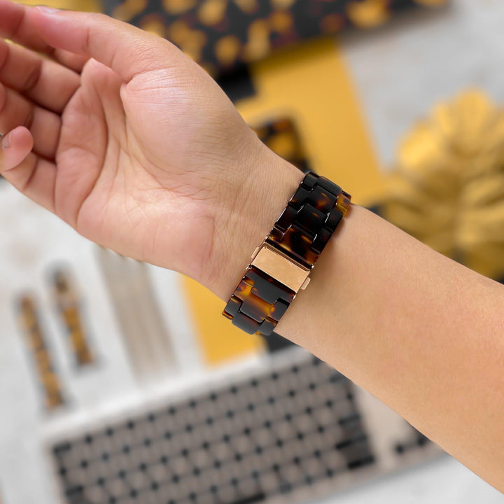 A close-up photo showcases a wrist wearing the Luxe Tortoiseshell Apple Watch Strap by Coconut Lane, featuring a stylish tortoiseshell resin design with a rose gold clasp, compatible with Apple Watch models. The background features a keyboard and other accessories in coordinating colors and patterns.