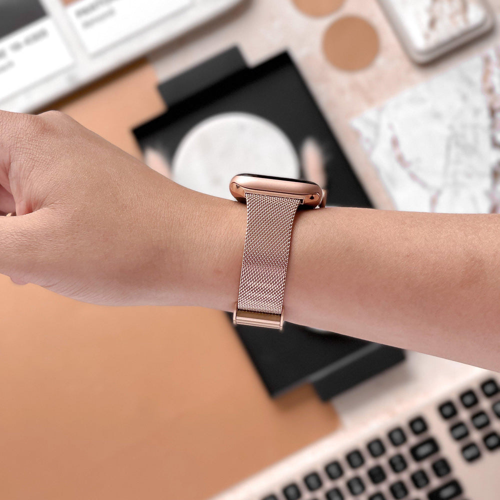 A person wears an Apple Watch with a Coconut Lane Milanese Apple Watch Strap - Rose Gold on their wrist. In the background, there is a keyboard, a marble-patterned notebook, and a partially visible phone on a desk. The overall scene has a mix of pastel and earthy tones.