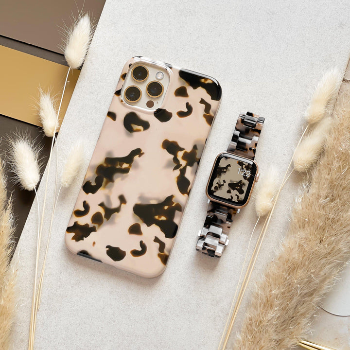 An Ivory Tort Phone Case from Coconut Lane, featuring a beige and black spotted pattern, is placed on a light grey surface alongside a matching smartwatch band with a similar animal print. Nearby, soft beige pampas grass and decorative elements in gold and brown tones add to the aesthetic. The smartwatch displays the time as 12:29.