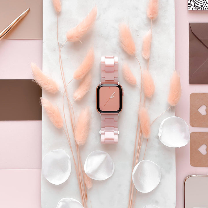 A square-faced smartwatch featuring the Luxe Pink Apple Watch Strap by Coconut Lane is showcased on a marble surface. Encircling the watch are delicate pink plants, white petals, and assorted stationery items such as notebooks and an envelope, all meticulously organized on a pink backdrop.