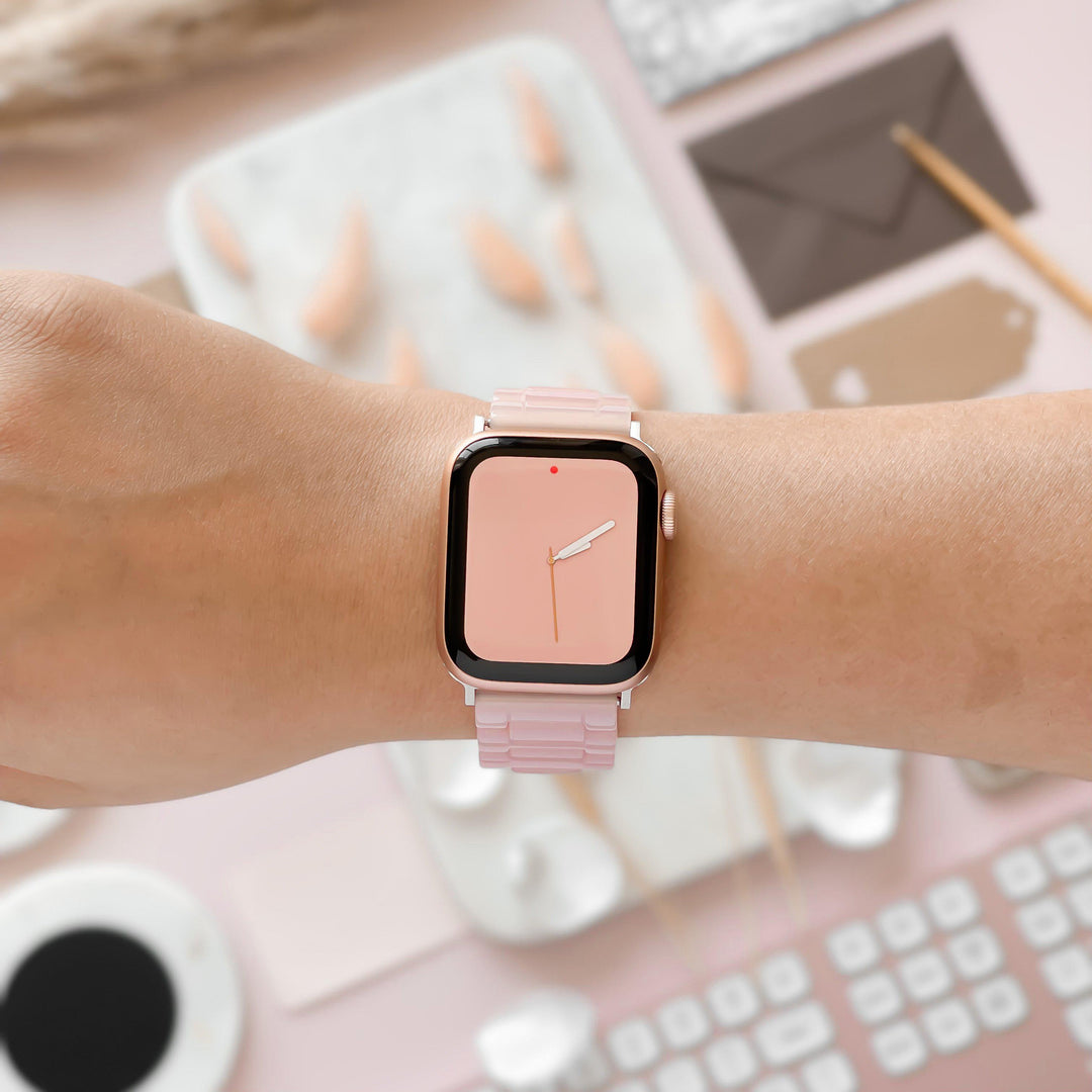 A close-up of a person's wrist featuring the Luxe Pink Apple Watch Strap by Coconut Lane, paired with a minimalistic pink clock face. The background displays a white keyboard, a coffee cup, and various stationery items in soft pastel tones.