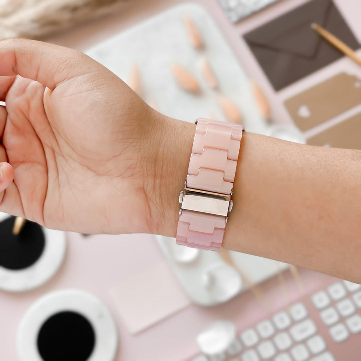 A close-up of a hand wearing the Luxe Pink Apple Watch Strap by Coconut Lane. The background features an assortment of items, including a keyboard, coffee cup, envelopes, and a pencil on a light pink surface. The look is enhanced with an adjustable strap that complements the chic setup perfectly.