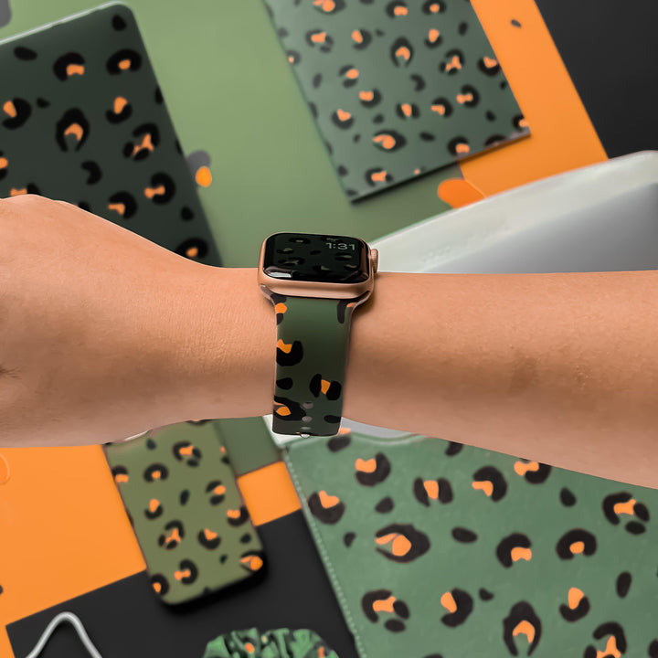 A hand adorned with Coconut Lane's Khaki Leopard Apple Watch Strap, showcasing a green leopard print band. The background is accessorized with tech items like phone cases, laptop covers, and earbud cases, all displaying the same animal print design against an orange and black backdrop.
