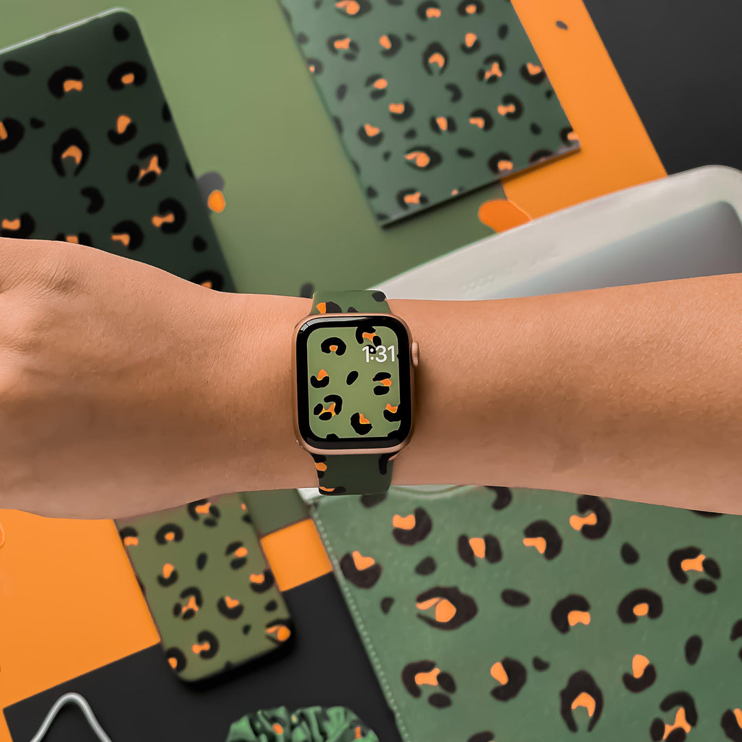 A hand wearing the Coconut Lane Khaki Leopard Apple Watch Strap with a matching leopard print watch face. The background displays an array of items, such as phone cases, notebooks, and a laptop cover, all boasting the same eye-catching khaki leopard print design.
