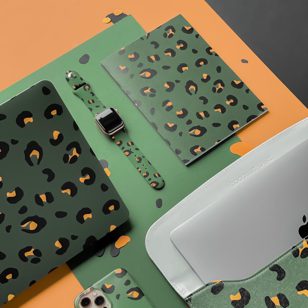 An assortment of green leopard print items, including a Coconut Lane Khaki Leopard Apple Watch Strap silicone smartwatch band, a laptop sleeve, a smartphone case, a journal, and a folder. The background features a combination of orange, green, and black surfaces.