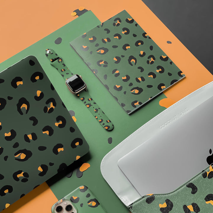 An assortment of green leopard print items, including a Coconut Lane Khaki Leopard Apple Watch Strap silicone smartwatch band, a laptop sleeve, a smartphone case, a journal, and a folder. The background features a combination of orange, green, and black surfaces.
