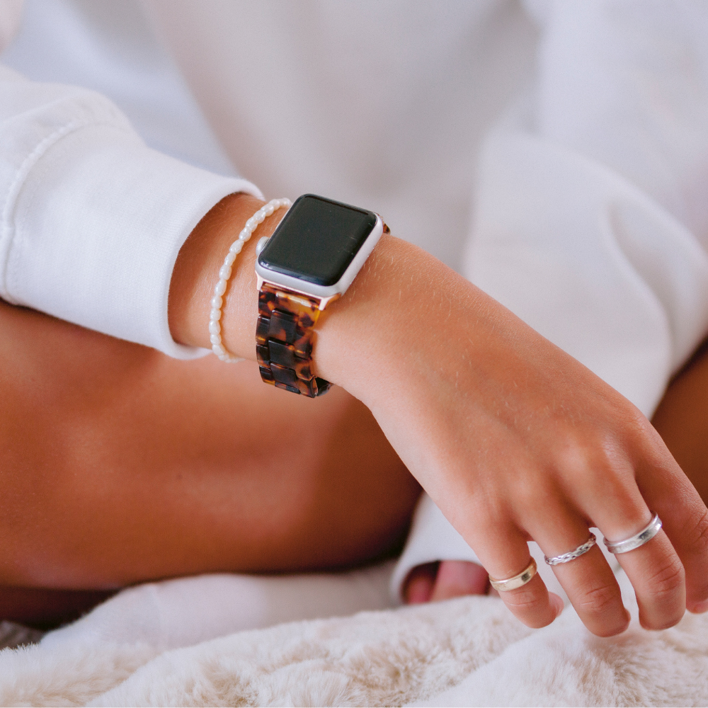 Close-up of a person's wrist adorned with the Luxe Tortoiseshell Apple Watch Strap by Coconut Lane. They are dressed in casual white attire, complemented by several bracelets and rings. The individual is seated with legs crossed on a soft surface, evoking a relaxed atmosphere.