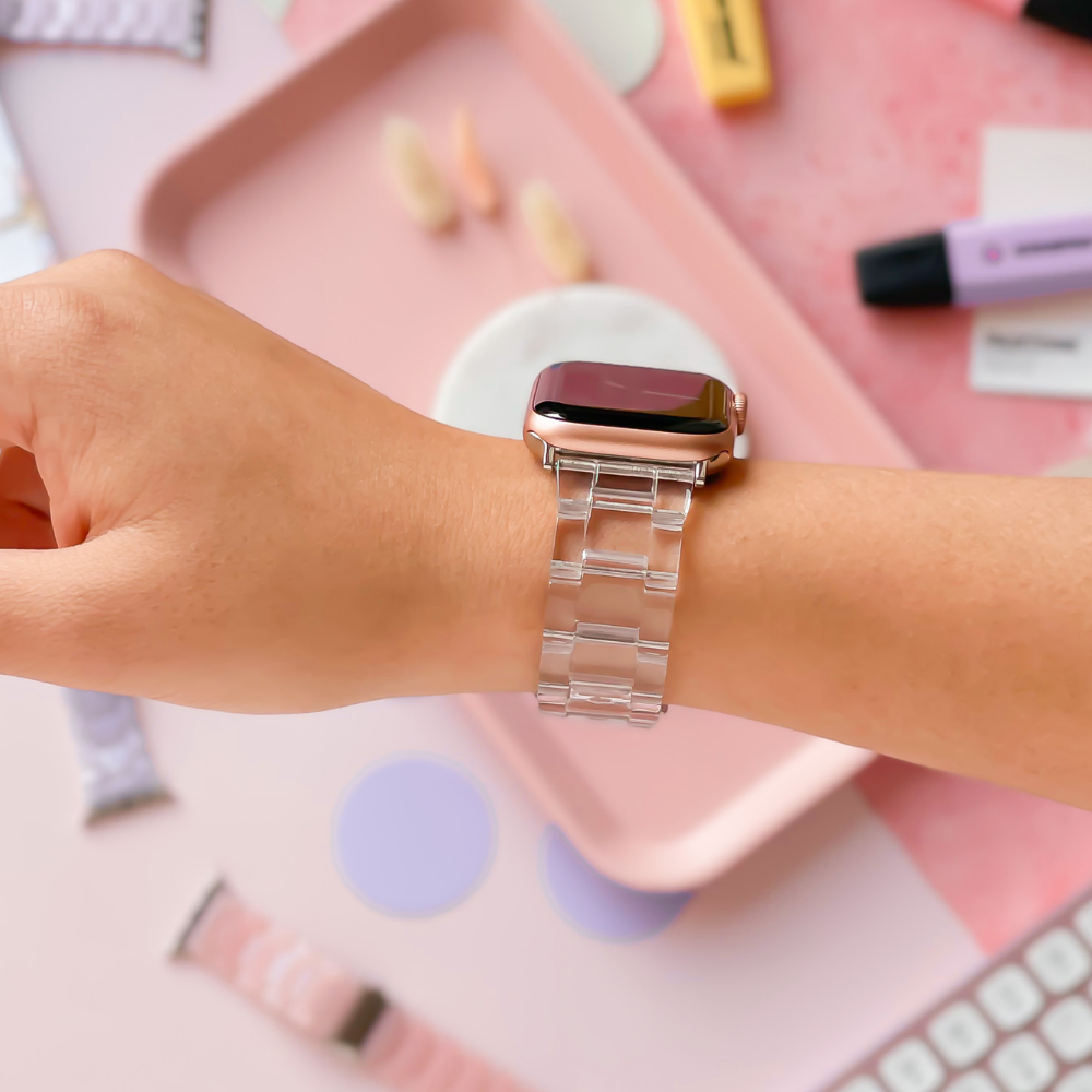 A person’s wrist adorned with the Luxe Transparent Apple Watch Strap by Coconut Lane elegantly complements the pastel-colored office supplies in the background. The scene includes a pink tray, several pens, and a keyboard, creating an organized and aesthetically pleasing workspace.