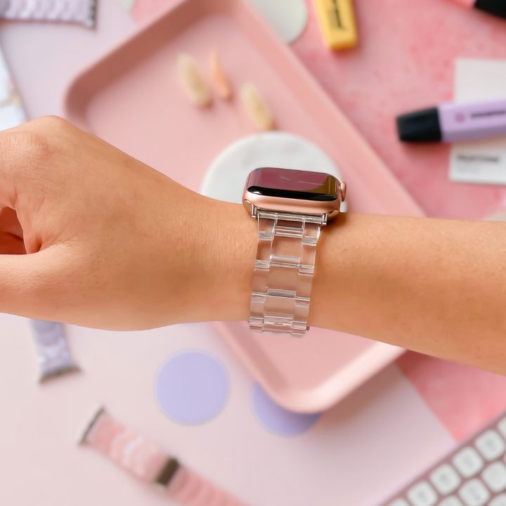 A person’s wrist adorned with the Luxe Transparent Apple Watch Strap by Coconut Lane elegantly complements the pastel-colored office supplies in the background. The scene includes a pink tray, several pens, and a keyboard, creating an organized and aesthetically pleasing workspace.