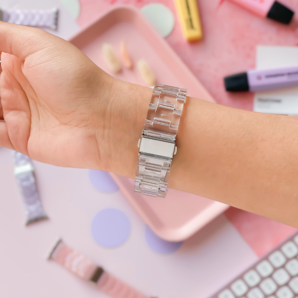 A hand is shown wearing a Coconut Lane Luxe Transparent Apple Watch Strap. The background features pastel-colored accessories, including tubes of skincare products, a keyboard, and a pink tray with scattered items. Various watch bands and circular paper pieces are also visible.
