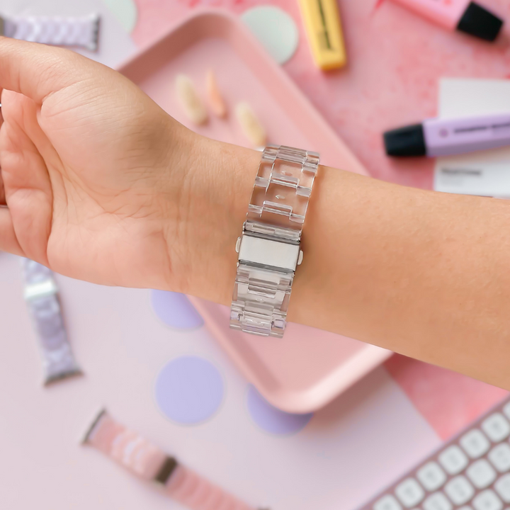 A hand is shown wearing a Coconut Lane Luxe Transparent Apple Watch Strap. The background features pastel-colored accessories, including tubes of skincare products, a keyboard, and a pink tray with scattered items. Various watch bands and circular paper pieces are also visible.