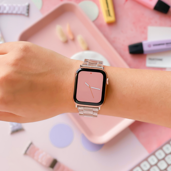 A person is shown wearing a smartwatch with a pink watch face and the Luxe Transparent Apple Watch Strap by Coconut Lane. The background showcases various pastel-colored stationery items, including highlighters, a keyboard, and a tray.