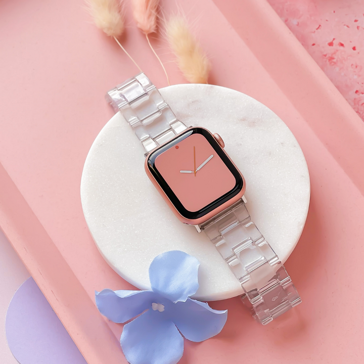 A Coconut Lane Luxe Transparent Apple Watch Strap paired with a smartwatch featuring a pink face is artfully presented on a round, white marble stand. The background showcases a pink surface adorned with decorative elements, including light blue flowers and beige dried plants.