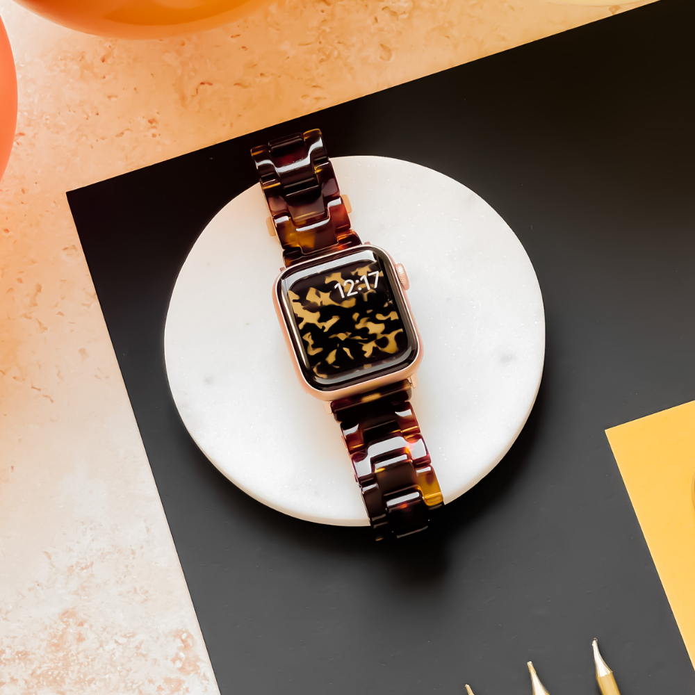 An Apple Watch adorned with the Luxe Tortoiseshell Apple Watch Strap by Coconut Lane rests on a round, white marble surface. The smartwatch screen displays the time "12:27" with an abstract black and gold design. The surrounding area features a black and beige background and a yellow paper.