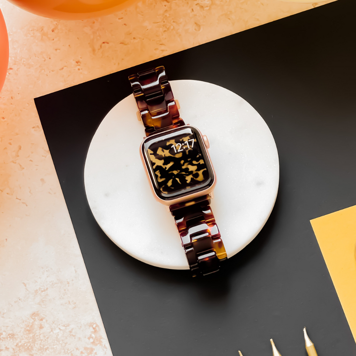 An Apple Watch adorned with the Luxe Tortoiseshell Apple Watch Strap by Coconut Lane rests on a round, white marble surface. The smartwatch screen displays the time "12:27" with an abstract black and gold design. The surrounding area features a black and beige background and a yellow paper.