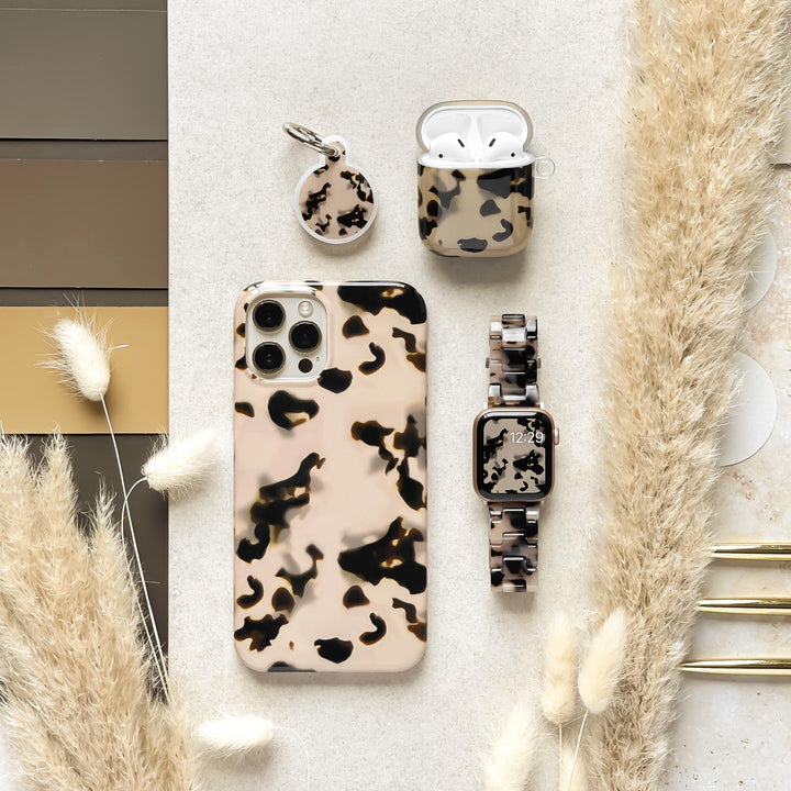 A flat lay image showcases a coordinated gadget accessory set in a beige and black animal print pattern. Items include an Ivory Tort Phone Case for iPhone 15 by Coconut Lane, a keychain, an AirPods case, and an Apple Watch with a matching band. Beige pampas grass decorates the edges of the composition, adding an elegant touch to the tortoiseshell aesthetic.
