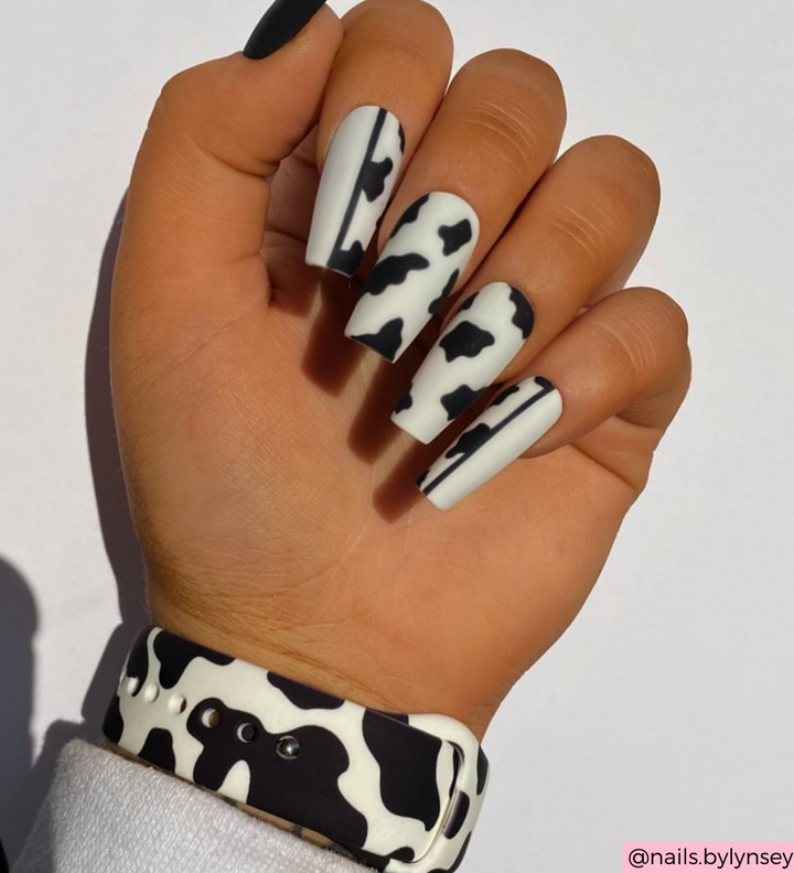 A hand with long, square-shaped nails showcases a cow print design in black and white. The wrist is decorated with Coconut Lane’s Cow Apple Watch Strap, creating a coordinated look. The neutral background highlights the nail art and the stylish Apple Watch accessory.